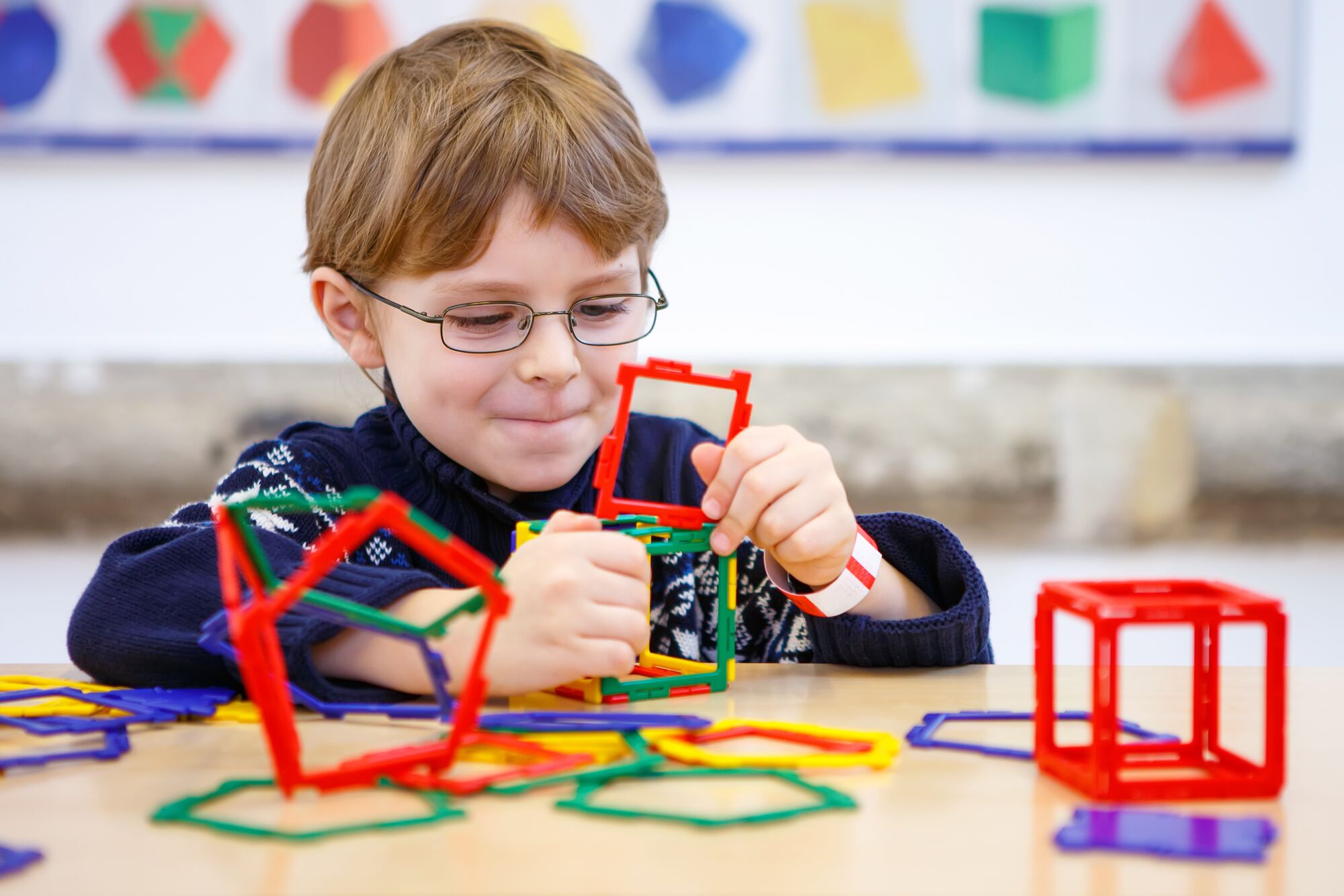 A boy with glasses builds a colorful geometric structure using plastic shapes on a table, focused and engaged in creative play.